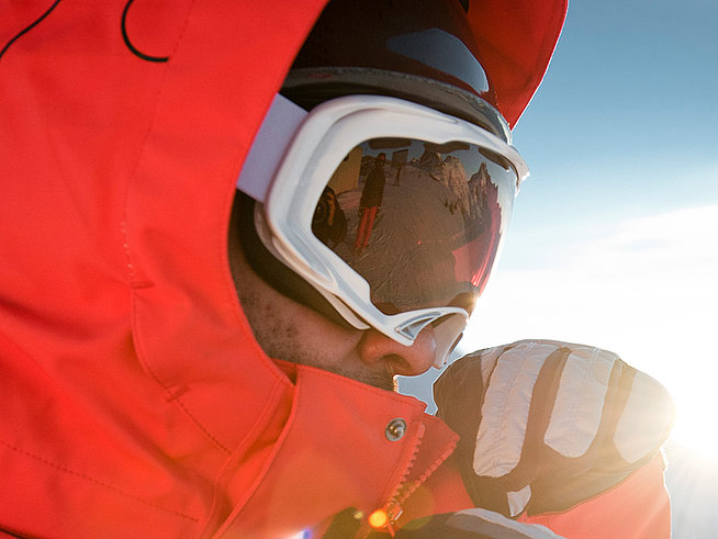 Skifahrer in roter Jacke mit Skibrille steht vor verschneiten Bergen im Sonnenlicht